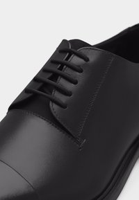 Μαύρο επίσημο παπούτσι από δέρμα με κλειστό κορδόνι, λείο άκρο και λεπτομερή ραφή σε απλό ανοιχτό φόντο.