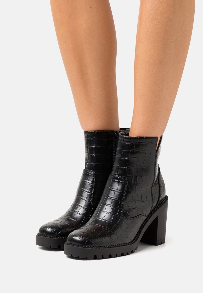 Jambes portant des bottines noires avec une texture crocodile, talons épais et semelles à crampons épaisses sur fond blanc.