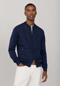 Hombre vistiendo una chaqueta bomber azul marino con cremallera sobre una camisa clara, con pantalones blancos y un cinturón marrón, posando frente a un fondo liso.