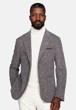 Man with short hair and beard wearing a gray textured blazer over a white cable knit turtleneck sweater and white pants.