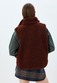 Brauner Fleece-Weste mit hohem Kragen, struktuierter Oberfläche; getragen über einer Jeansjacke und einem hellen karierten Rock. Sauberer, minimalistischer Hintergrund.