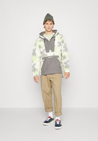 Sweatshirt leve e estampado com cores cinza e verde néon, fecho frontal em zíper, capuz ajustável e bolso canguru. Calças bege e ténis.