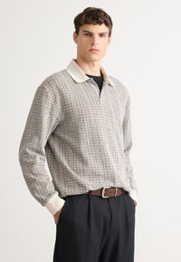 Young man wearing a patterned long-sleeve collared shirt tucked into black pants with a brown belt, standing against a plain background.