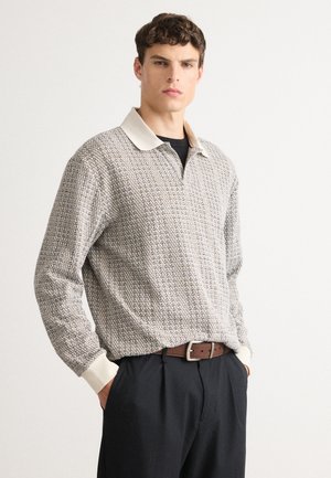 Young man wearing a patterned long-sleeve collared shirt tucked into black pants with a brown belt, standing against a plain background.