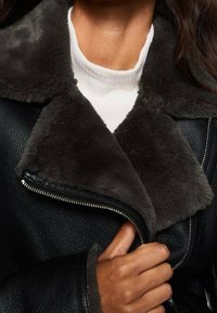 Persoon in een zwart leren jack met een donkergrijze bontkraag, gedeeltelijk open, over een witte geribde coltrui.