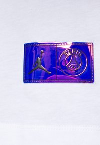 Vit tygtröja med en rektangulär, iriserande patch med en svart Air Jordan-logotyp och texten "Paris Saint Germain".