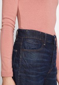 Jeans de mezclilla con un lavado azul oscuro, con costuras visibles, bolsillos delanteros y una cintura alta, combinados con una blusa de manga larga rosa claro.
