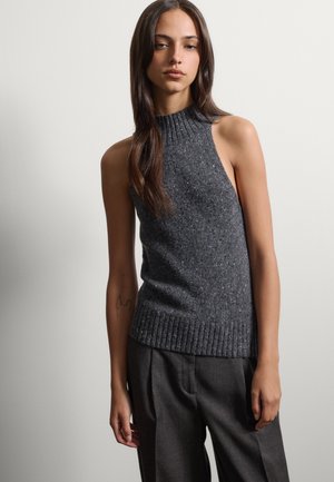 Jeune femme aux longs cheveux foncés portant un haut en maille gris foncé sans manches et un pantalon gris foncé sur un fond clair uni.