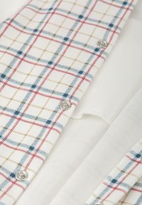 Tela de camisa a cuadros en blanco, con líneas azules, rojas y marrones, con botones a presión plateados y suave algodón blanco por debajo.