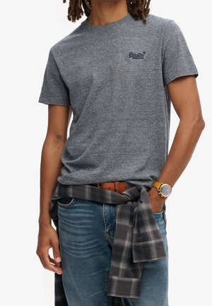 Grå kortærmet t-shirt med rund hals og et lille logo på brystet. Båret med blå jeans og et mørkt ternet flannel-skjorte bundet om livet.