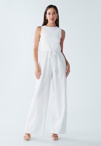 Witte mouwloze jumpsuit met een getextureerd lijfje en een koord taille. Uitlopende wijde pijpen en lichtgewicht stof, gecombineerd met witte sandalen.