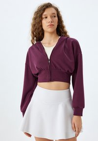 Sudadera corta con cremallera en color púrpura y mangas largas, hecha de una tela suave. Combinada con una falda blanca. Diseño simple sin patrones visibles.
