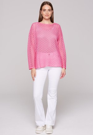 Pink open-knit pullover with diamond pattern, long sleeves, and a relaxed fit, paired with white flared pants and light-colored sneakers.