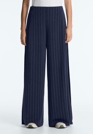 Woman wearing navy blue wide-leg pants with thin white vertical stripes and white sneakers, standing against a plain background.