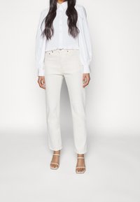 Hvid skjorte med pufærmer, parret med lyse beige lige bukser og sandaler med klare stropper. Glat stof og klassisk design.