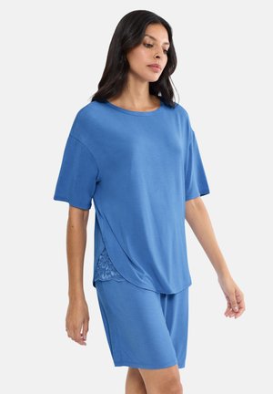 Kobieta ubrana w luźną, niebieską bluzkę z krótkim rękawem i zaokrąglonym dołem, z koronkowymi zdobieniami, w połączeniu z dopasowanymi niebieskimi szortami sięgającymi do kolan.