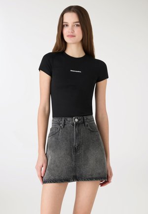 Giovane donna che indossa una t-shirt nera aderente con piccolo testo bianco e gonna di jeans nera slavata a vita alta, in piedi davanti a uno sfondo semplice.