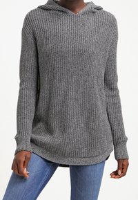 Grijze gebreide hoodie trui met een textuurpatroon, lange mouwen en een gebogen zoom. Heeft een capuchon en een relaxte pasvorm. Gecombineerd met blauwe jeans.