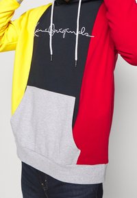 Sudadera con capucha que presenta un diseño a bloques de color en amarillo, rojo, gris y negro, con un bolsillo frontal y cordones blancos.