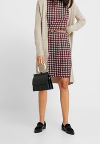 Robe bordeaux à motifs avec des designs géométriques blancs, cardigan beige, sac à main noir avec une anse circulaire, et mocassins noirs.
