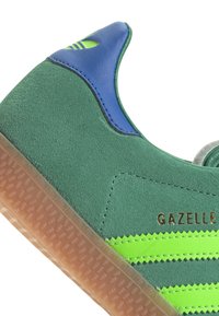 Zelené semišové tenisky s modrými akcentmi, tromi zelenými pruhmi a gumovou podrážkou. Obsahujú zlaté logo "GAZELLE".