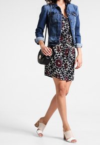 Džínová bunda přes vzorované šaty s geometrickými tvary v tmavě fialové a bílé. Bílé otevřené muly na podpatku z korku a černá crossbody kabelka.