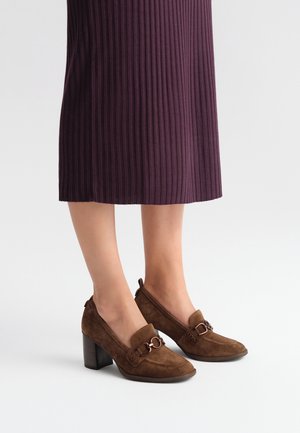 Scarpe con tacco in suede marrone con punta arrotondata, con un'accattivante fibbia metallica decorativa e un tacco robusto, abbinate a una gonna plissettata color prugna.
