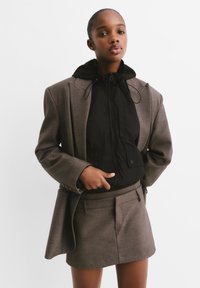 Brown tailored blazer over a black zip-up jacket with a hood, worn with a matching short skirt featuring a flat front and side pockets.