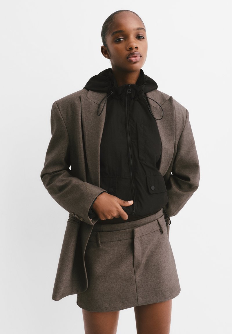 Brown tailored blazer over a black zip-up jacket with a hood, worn with a matching short skirt featuring a flat front and side pockets.