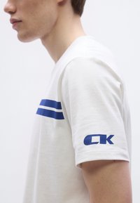 Bela bombažna majica z modrimi horizontalnimi črtami čez prsni koš in modrim logotipom na rokavu. Enostaven, priložnosten dizajn.