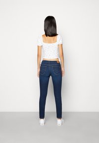 Vaqueros ajustados de mezclilla azul oscuro con bolsillos traseros y una etiqueta de cuero. Combinados con una blusa blanca con estampado floral y zapatos blancos.