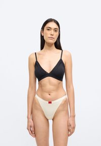 Bralette negro con escote en V y tirantes delgados, combinado con un tanga crema que lleva una etiqueta de logo rojo en la cintura. Textura de tela suave.