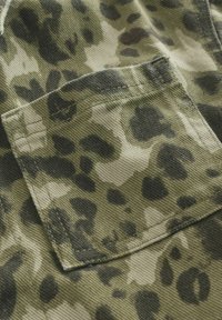 Tissu camouflage vert avec une poche proéminente, présentant une texture et des taches irrégulières noires et vert foncé.