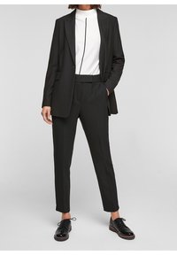 Svart skräddarsydd kavaj över en vit topp med ett vertikalt svart streck, kombinerat med svarta byxor och svarta finskor. Slät textur och åtsittande design.