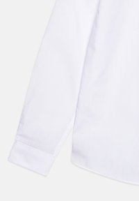Manche longue de chemise blanche avec poignet boutonné, tissu lisse et léger détail de couture près du poignet et de la couture latérale.