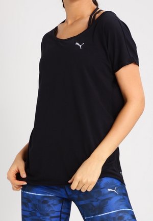 Frau trägt schwarzes Puma-T-Shirt mit kurzen Ärmeln und blaue gemusterte Puma-Leggings, steht mit einer Hand, die den Saum des Shirts hält.