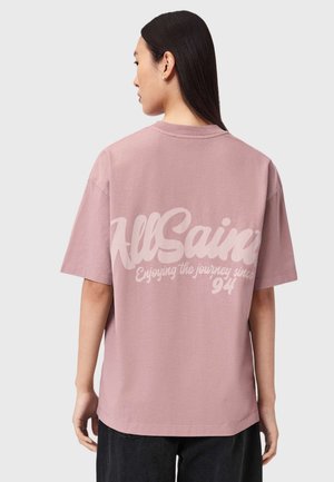 Kvinde med langt mørkt hår iført en løs støvet lyserød T-shirt med teksten "AllSaints Enjoying the journey since '94" på ryggen.