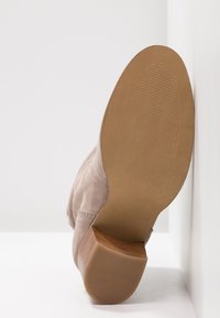 Bottine en daim beige clair posée sur le dos, montrant une semelle lisse de couleur fauve avec une surface texturée antidérapante et un petit talon empilé en bois.