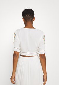 Top corto bianco con ricami floreali sulle maniche, orlo arricciato e tessuto testurizzato. Vista posteriore, che mostra uno stile semplice ed elegante.