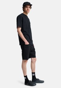 Sort bomulds t-shirt, sorte denim shorts, sorte sokker med logo, og mørke klipklapper; stående positur, sidebillede, neutral baggrund.