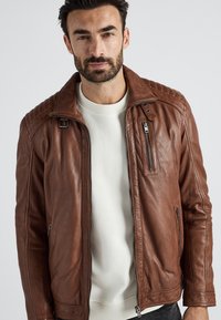 Bruine leren jas met een licht gestructureerde afwerking, voorzien van een staande kraag, ritszakken en gestikte schouderaccenten.