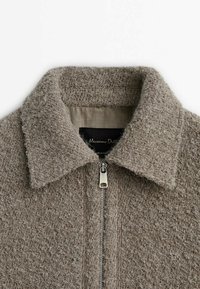 Brun strukturerad ulljacka med krage och silverfärgad dragkedja, visar övre framsida och insidan av etiketten.