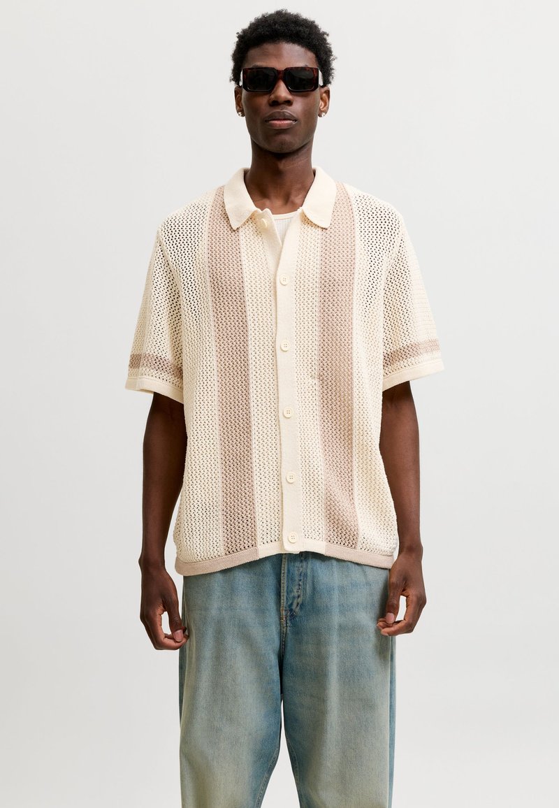 Homme portant des lunettes de soleil foncées, une chemise en maille beige et crème à manches courtes boutonnée, et un jean bleu ample, debout devant un fond uni.