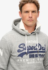 Felpa grigia con cappuccio e lacci bianchi, caratterizzata da un grande logo e testo 'SuperDry' blu navy sul davanti. Tessuto morbido e vestibilità casual.