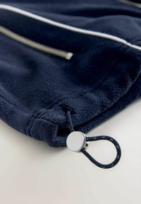 Tumesinist fliisriie kangas hõbedase tõmblukuga ja reguleeritava ümara nöörilukustiga, mis kinnitub tõmmissisaldise aasale.