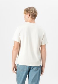 T-shirt à manches courtes et col rond en coton blanc cassé. Coupe droite avec des coutures minimales. Porté avec un jeans bleu clair, offrant un look décontracté.