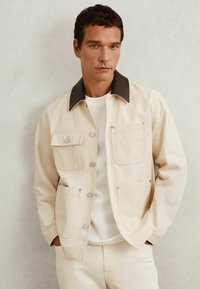 Beige, tekstureret jakke med to forlommer og en mørkebrun krave. Fastgjort med metalknapper, båret over en hvid skjorte.