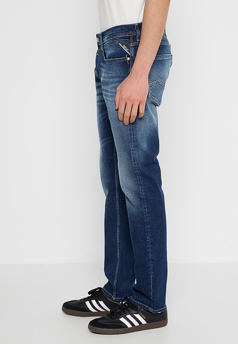 Mann in blauen, ausgewaschenen Jeans und schwarzen Adidas Samba Sneakers mit weißen Streifen, stehend vor weißem Hintergrund.