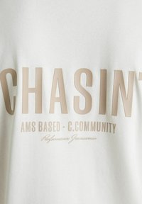Bela tkanina z bež besedilom "CHASIN'", manjše besedilo spodaj pravi "AMS BASED - C.COMMUNITY" in pisano "Performance Jeanswear."