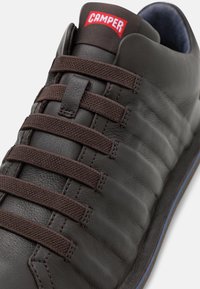Ciemnobrązowe skórzane trampki o teksturowanej powierzchni, z płaskimi brązowymi sznurówkami i czerwoną naszywką "CAMPER" na języku.